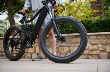 A person stands beside a black fat-tire electric bike on a paved walkway.