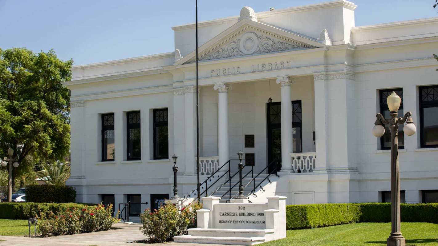 A white public library building with steps, lampposts, and surrounding greenery.