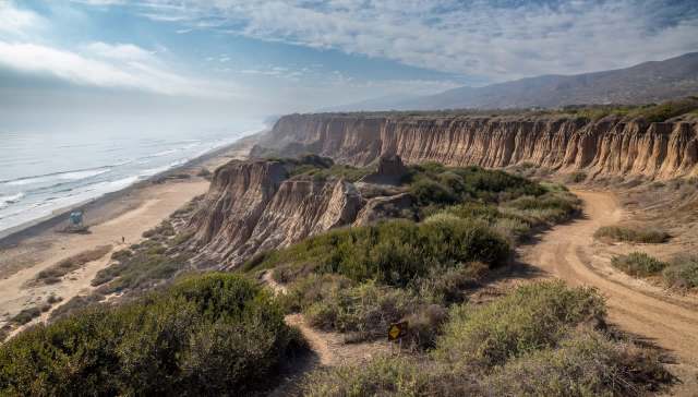 A coastal landscape featuring cliffs, a sandy beach, and gentle waves under a cloudy sky.