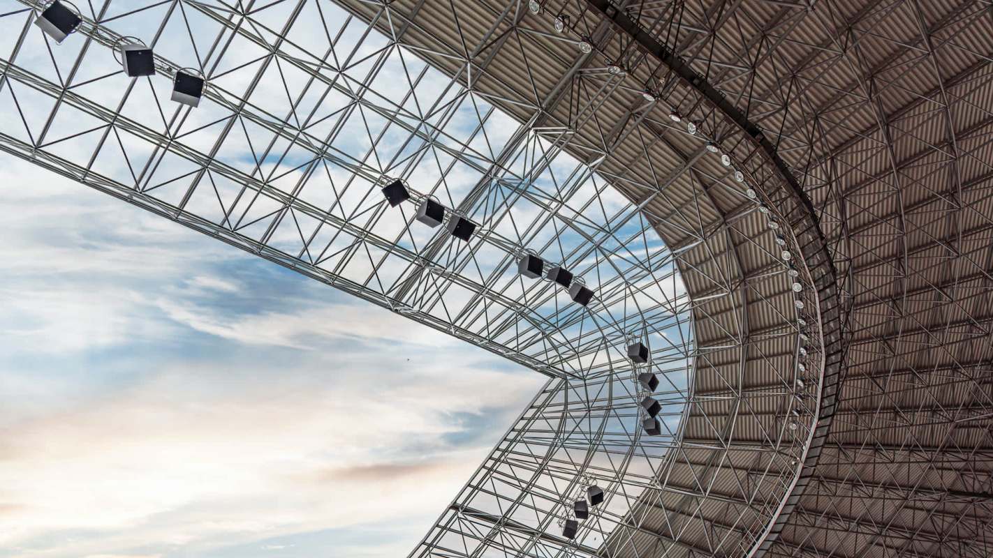 A geometric steel structure with a curved roof and hanging lights against a cloudy sky.
