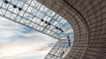 A geometric steel structure with a curved roof and hanging lights against a cloudy sky.
