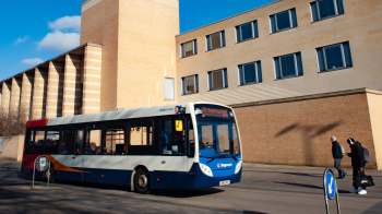 A bus drives past a building while two people walk on the sidewalk nearby.