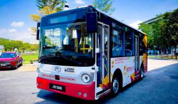 An autonomous electric bus parked on a street surrounded by trees and a red car.