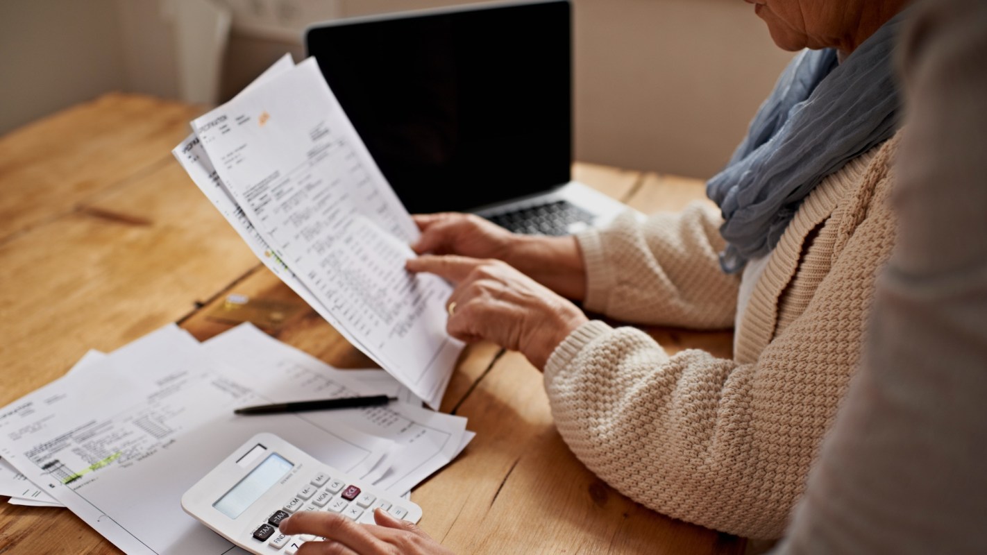 A person calculates expenses while reviewing documents and using a calculator at a wooden table.