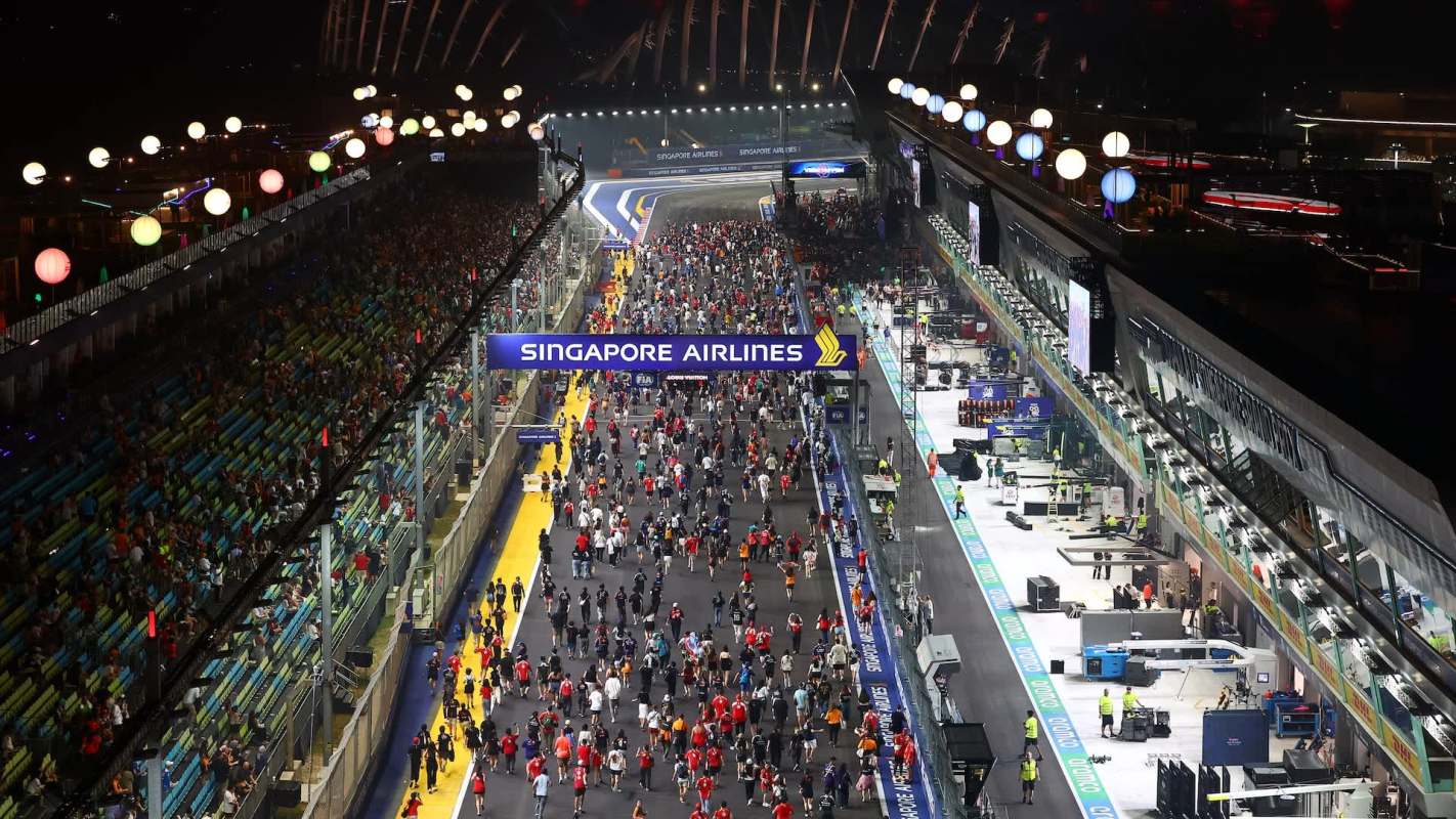 A crowd of people at the Formula 1 Singapore Grand Prix walks along a racetrack beside automotive team garages at night.