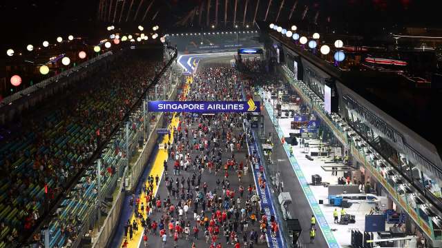 A crowd of people at the Formula 1 Singapore Grand Prix walks along a racetrack beside automotive team garages at night.