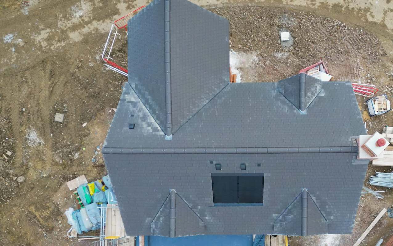 An aerial view of a house under construction with a dark sloped roof and surrounding construction materials.