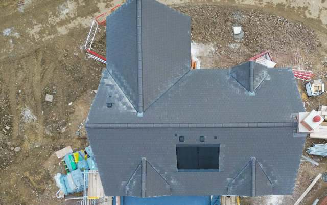 An aerial view of a house under construction with a dark sloped roof and surrounding construction materials.