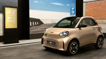 A small golden car by Smart displayed in an indoor setting with promotional signage in the background.