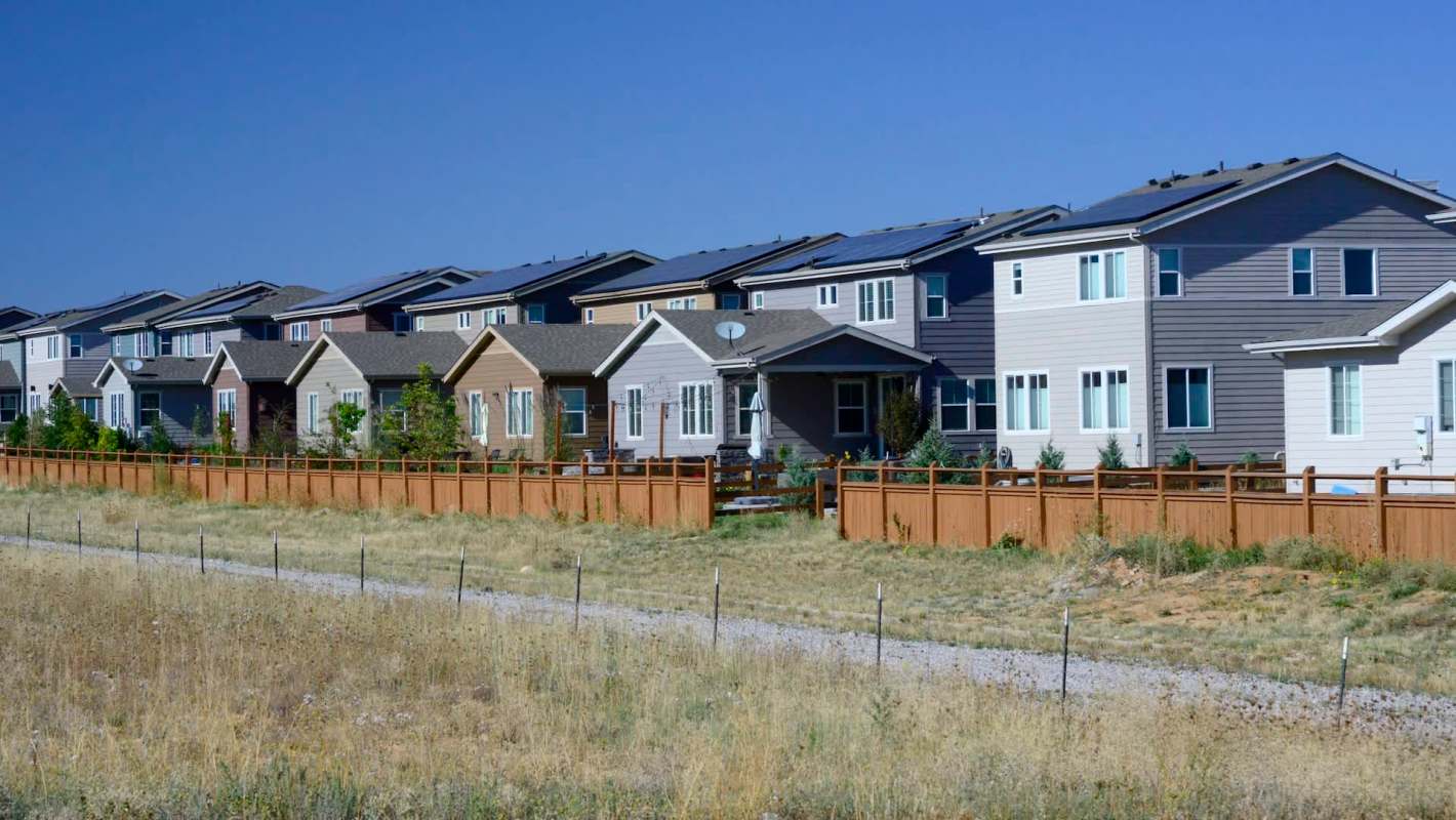 A row of residential houses with fences and solar panels, set against a clear blue sky.