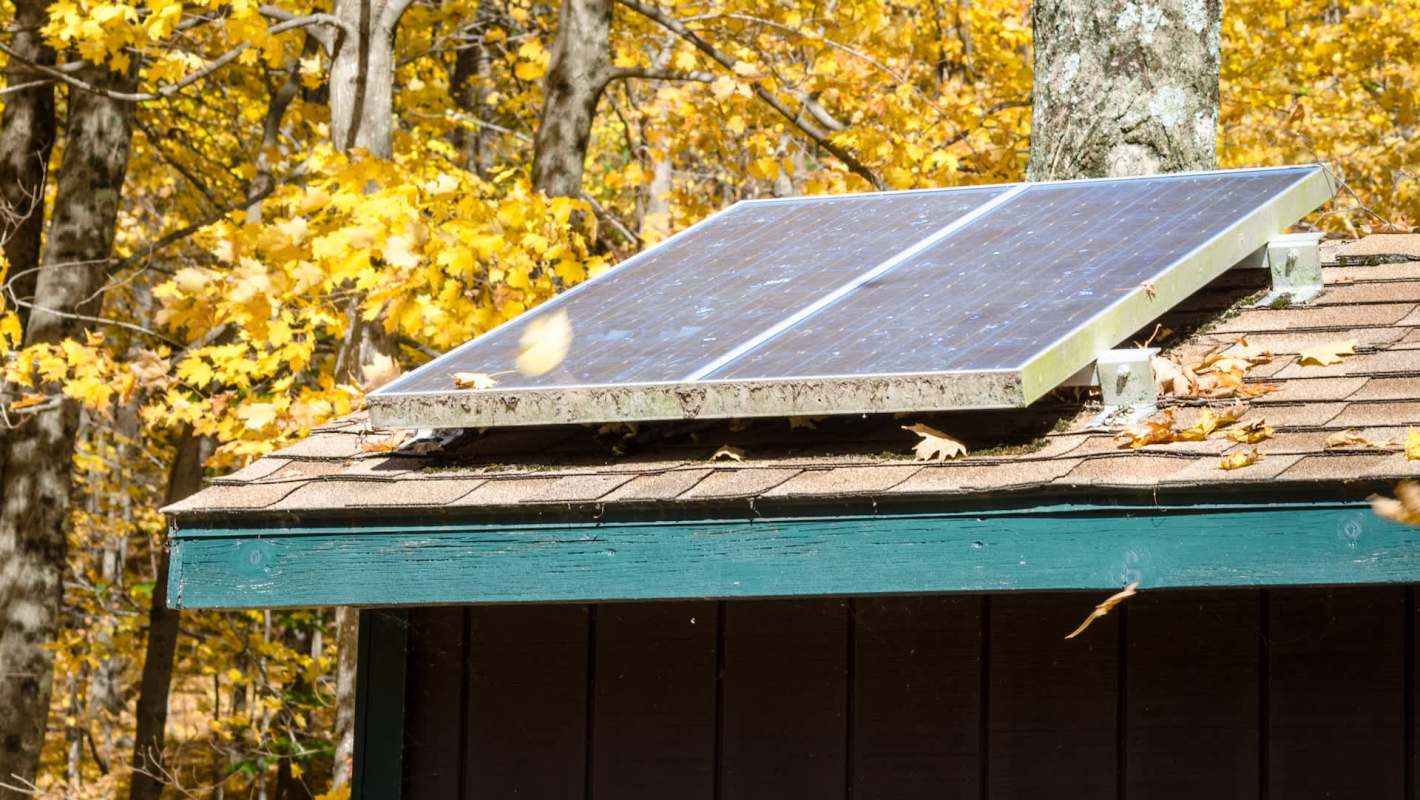 A solar panel installed on the roof of a cabin surrounded by autumn foliage.