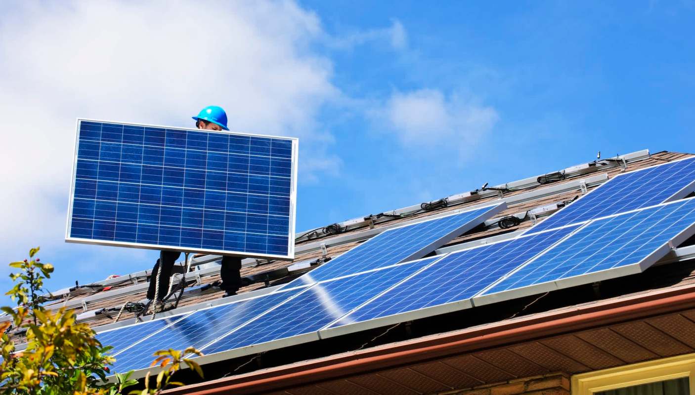 A person installs a solar panel on a roof covered with solar panels under a blue sky.