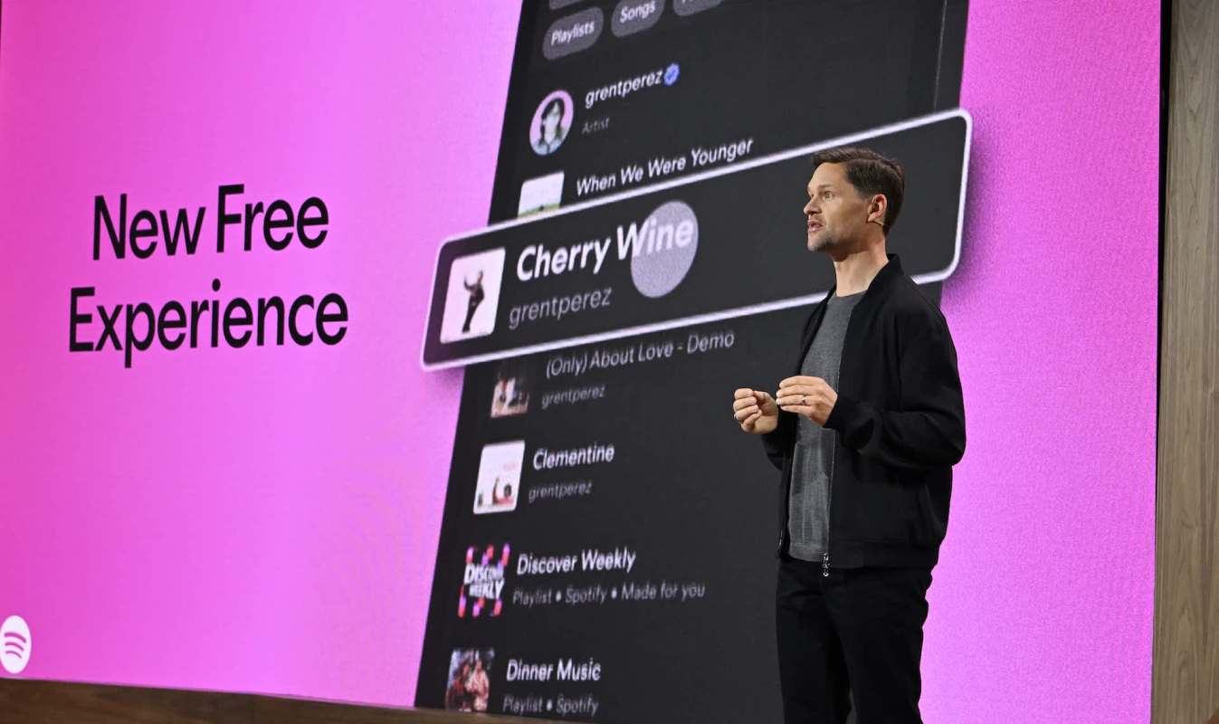 A man speaks on stage beside a screen displaying a Spotify interface on a smartphone.