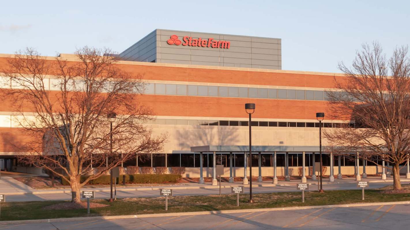 A brick building with large windows and a sign for State Farm, alongside a parking area marked for visitors.
