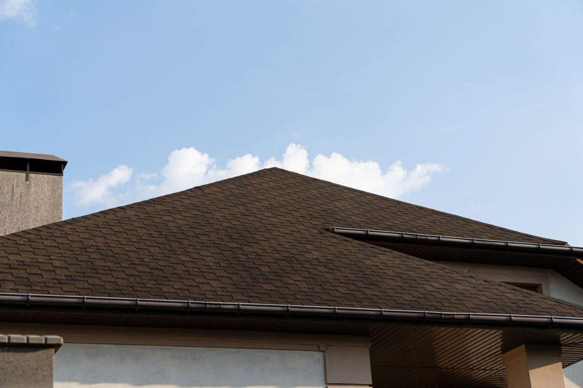 A steep brown shingle roof under a clear blue sky with a few clouds.