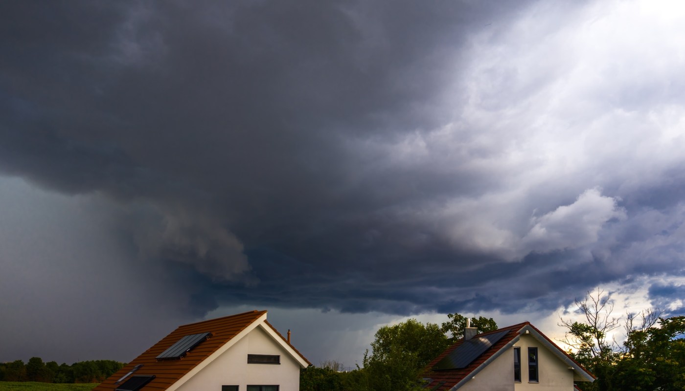 Dark storm clouds loom over two houses with pitched roofs and solar panels, surrounded by trees.