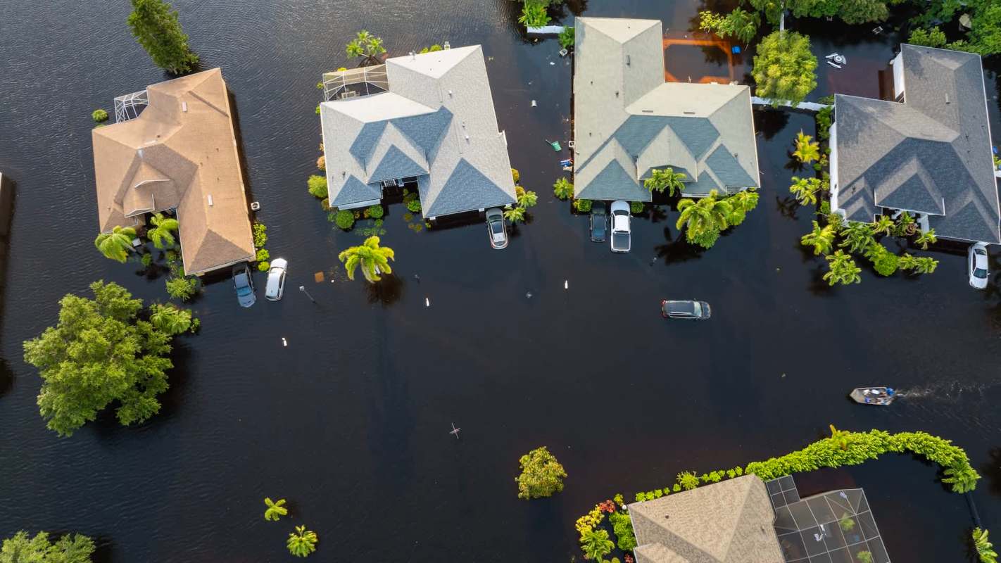 Several houses surrounded by floodwater, with some vehicles partially submerged and a boat navigating the area.