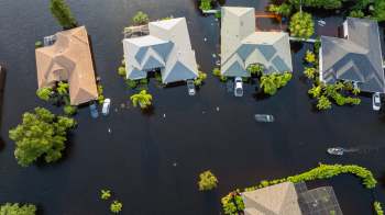 Several houses surrounded by floodwater, with some vehicles partially submerged and a boat navigating the area.