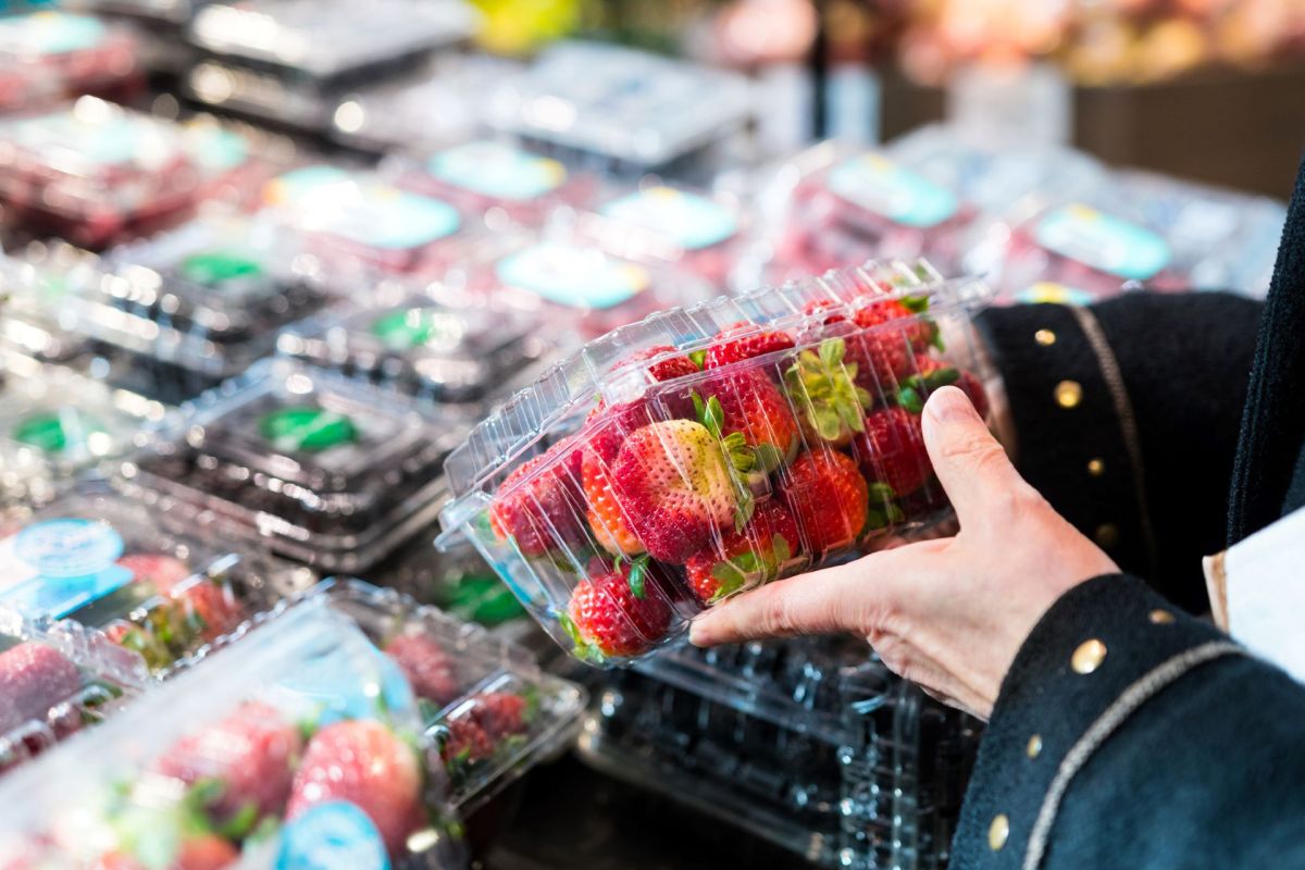 A person holding a container of strawberries in a market with various fruit containers in the background.