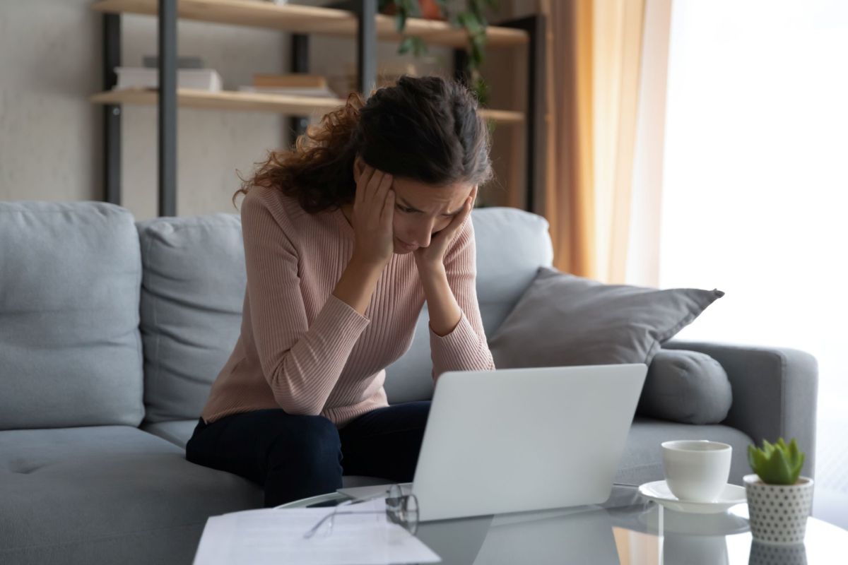 A stressed woman sitting on a couch, looking at a laptop.