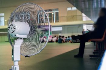 A standing electric fan in a spacious indoor area with students sitting in the background.