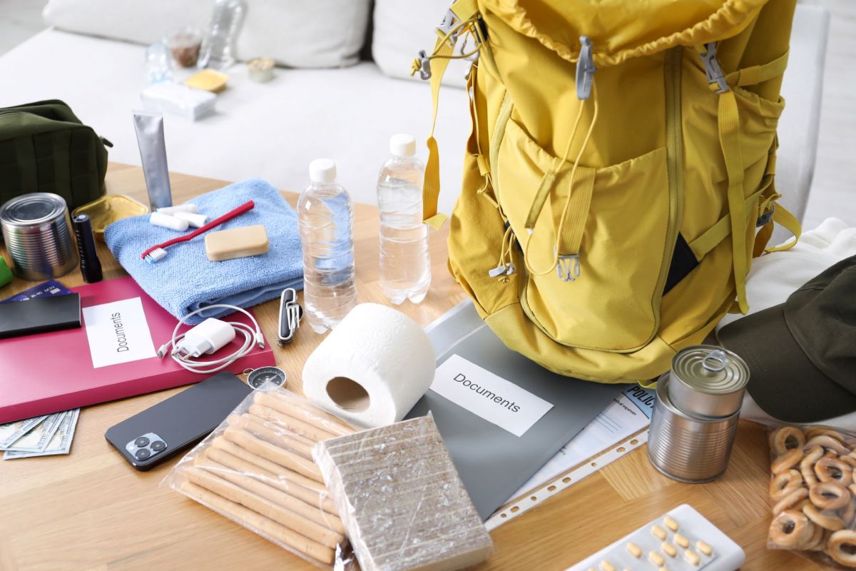 A yellow backpack surrounded by various travel items on a wooden table, including water bottles and snacks.