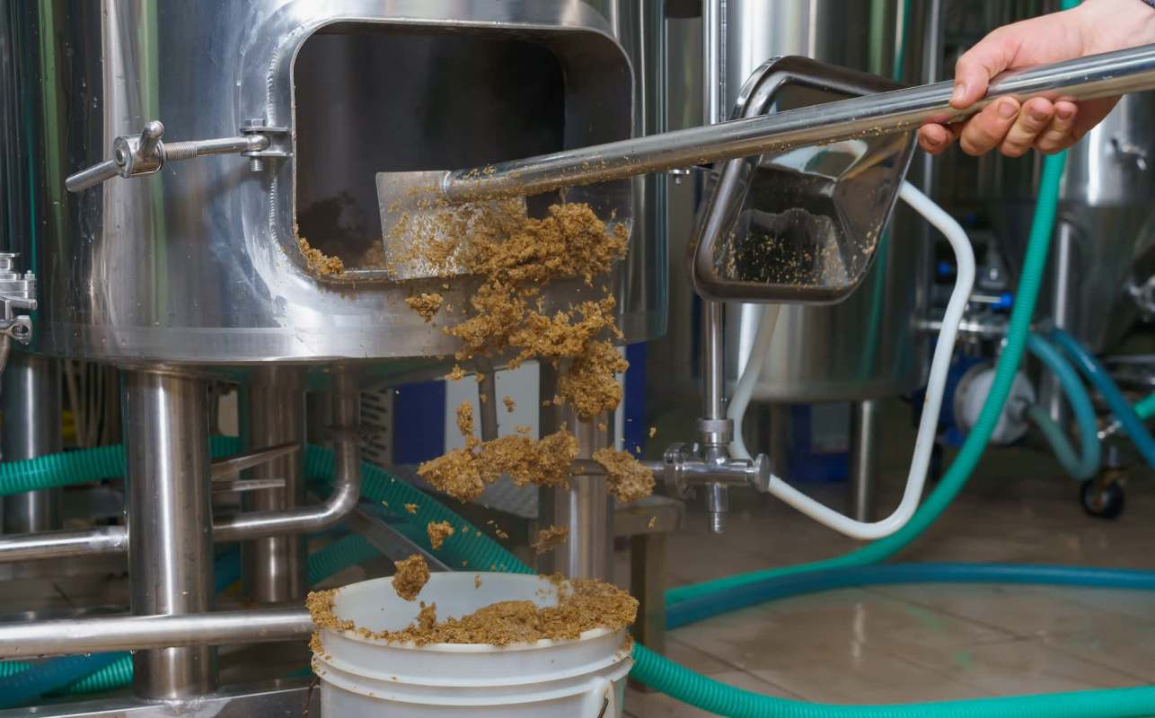A person uses a shovel to transfer spent grain from a brewing vessel into a bucket.