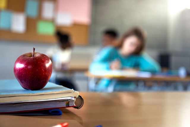 An apple sits on top of stacked notebooks in a classroom with students in the background.