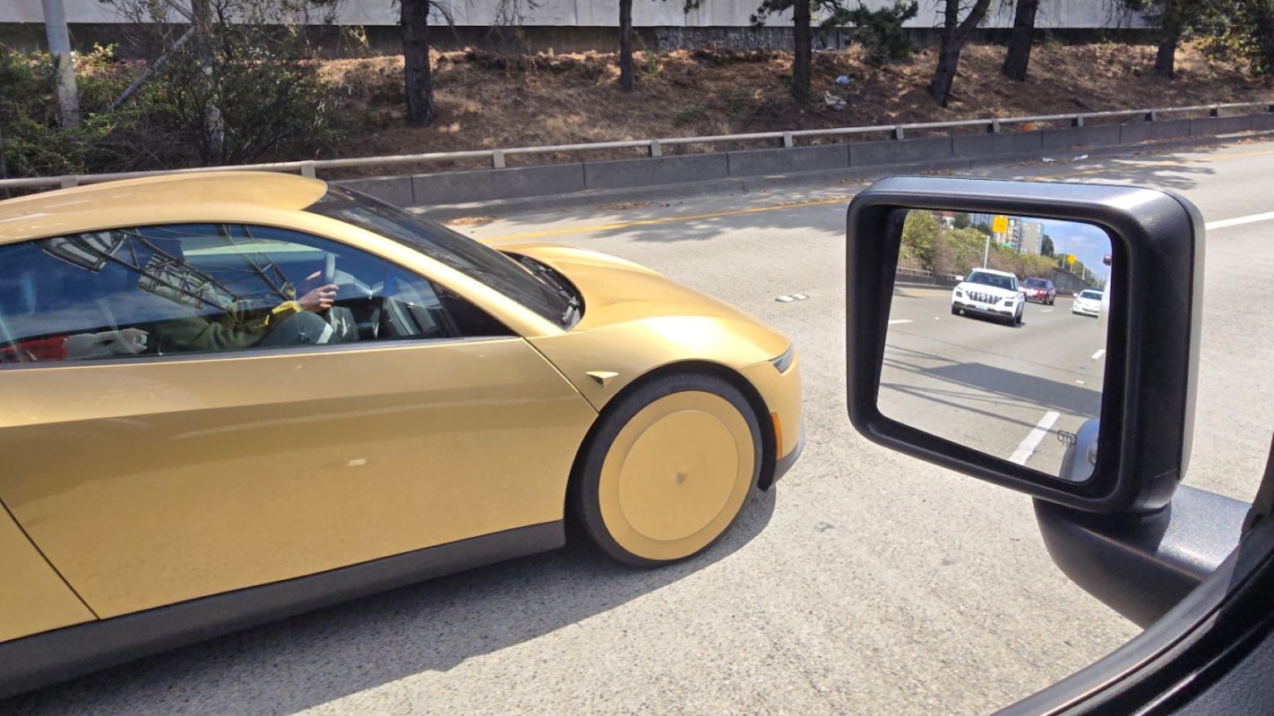 A gold electric car driving on the highway with other vehicles visible in the side mirror.