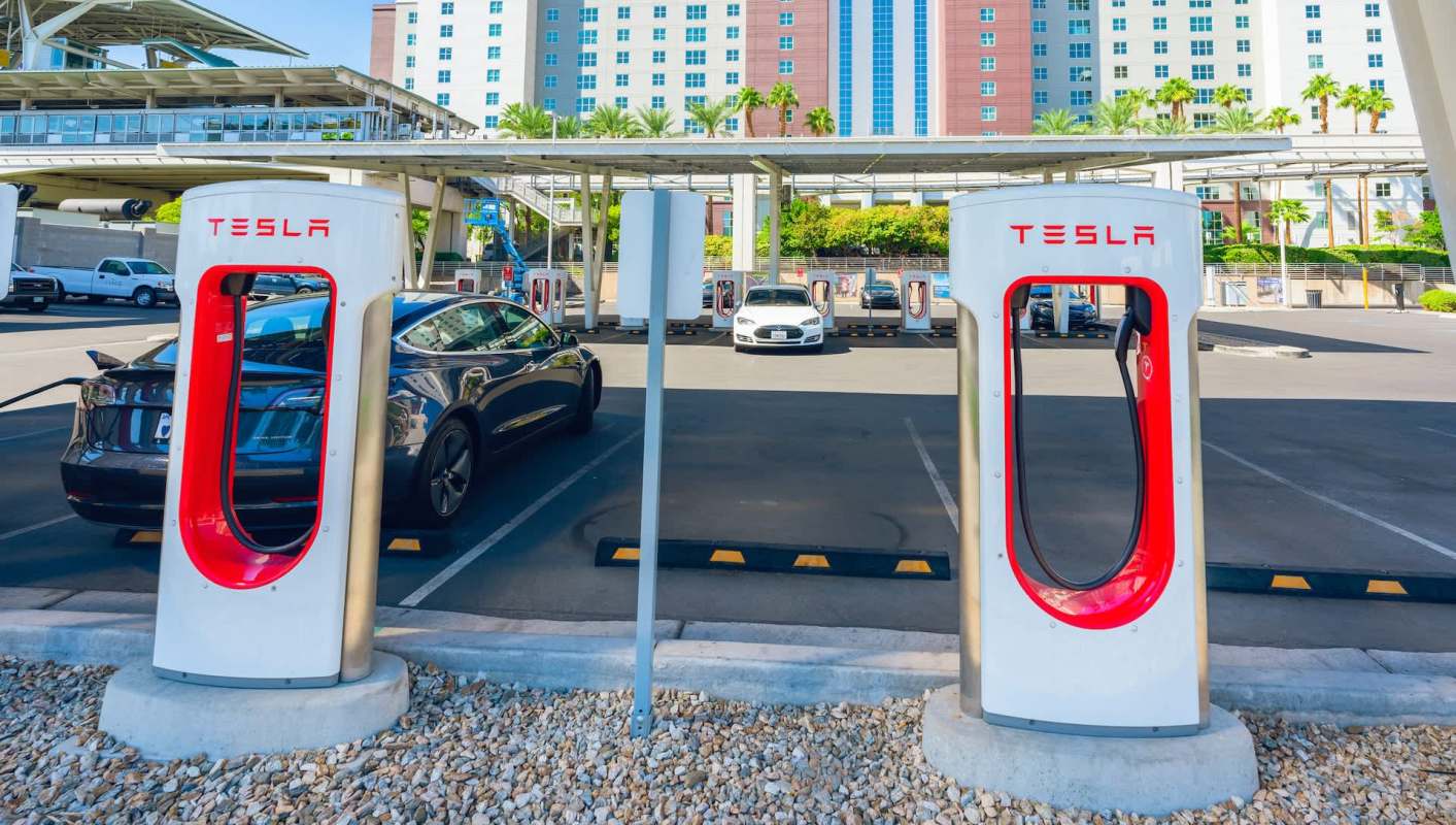 Tesla charging stations in a parking lot with several electric cars and a building in the background.