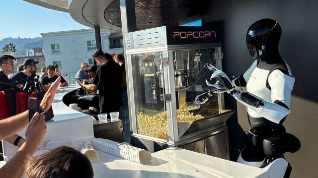 A Tesla Optimus robot hands popcorn to a customer at a food counter with a group of people in the background.