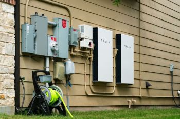 Two Tesla home battery units mounted on the side of a house near electrical equipment and a garden hose reel.