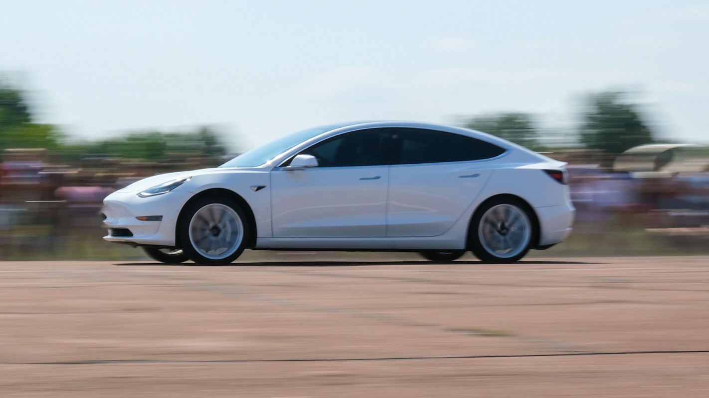 A white car is driving rapidly on a paved surface with spectators in the background.