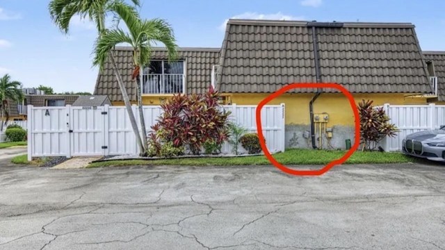 A building with a yellow wall, pipes, and a white fence, surrounded by plants and a parked car.