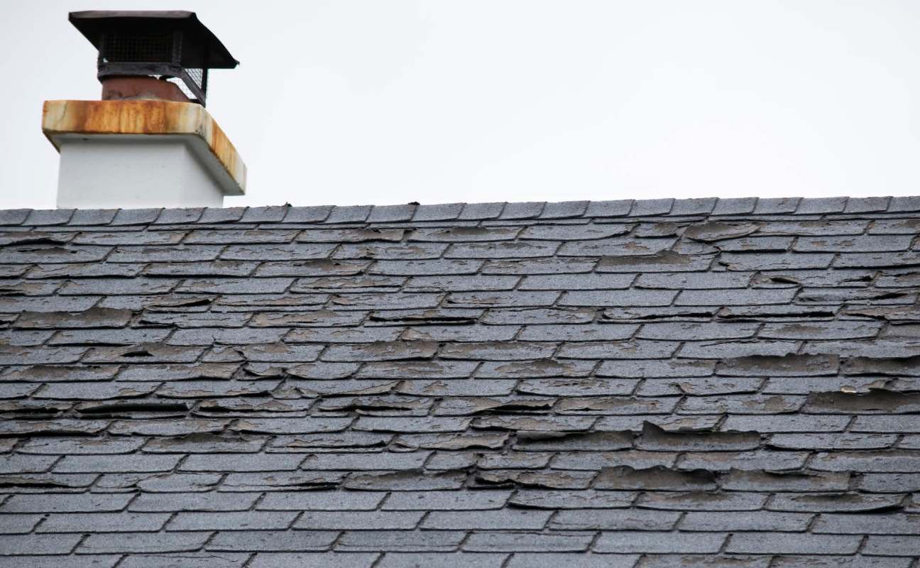 A roof with damaged and peeling shingles, featuring a chimney with a rusted cap.