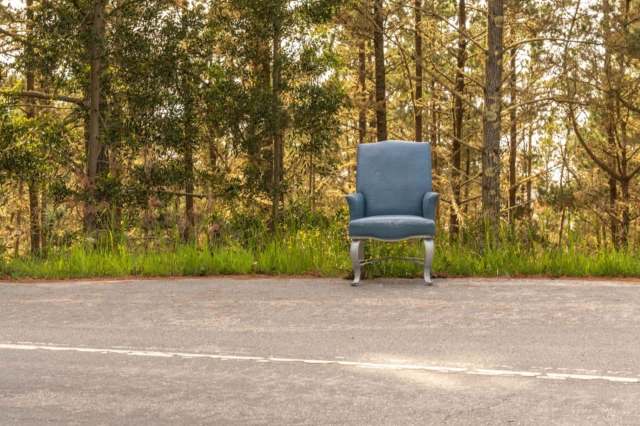 An armchair is placed on the side of a road next to trees and grass.