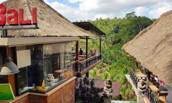 A restaurant with a thatched roof and a Bali sign, overlooking a lush green landscape, with multiple diners seated outside.