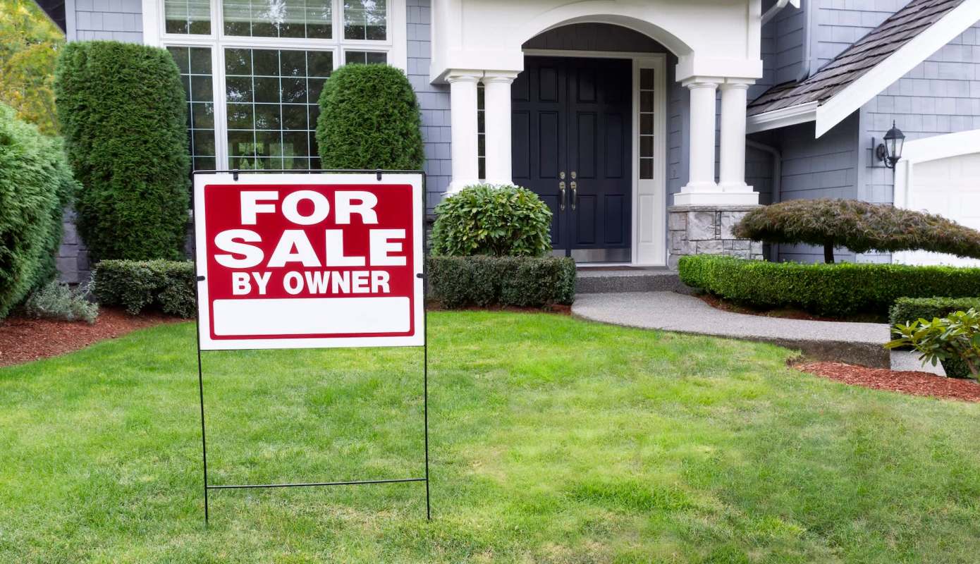 A "For Sale By Owner" sign in a well-maintained front yard of a house.