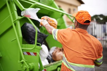 A worker in an orange uniform dumps trash into a green garbage truck.