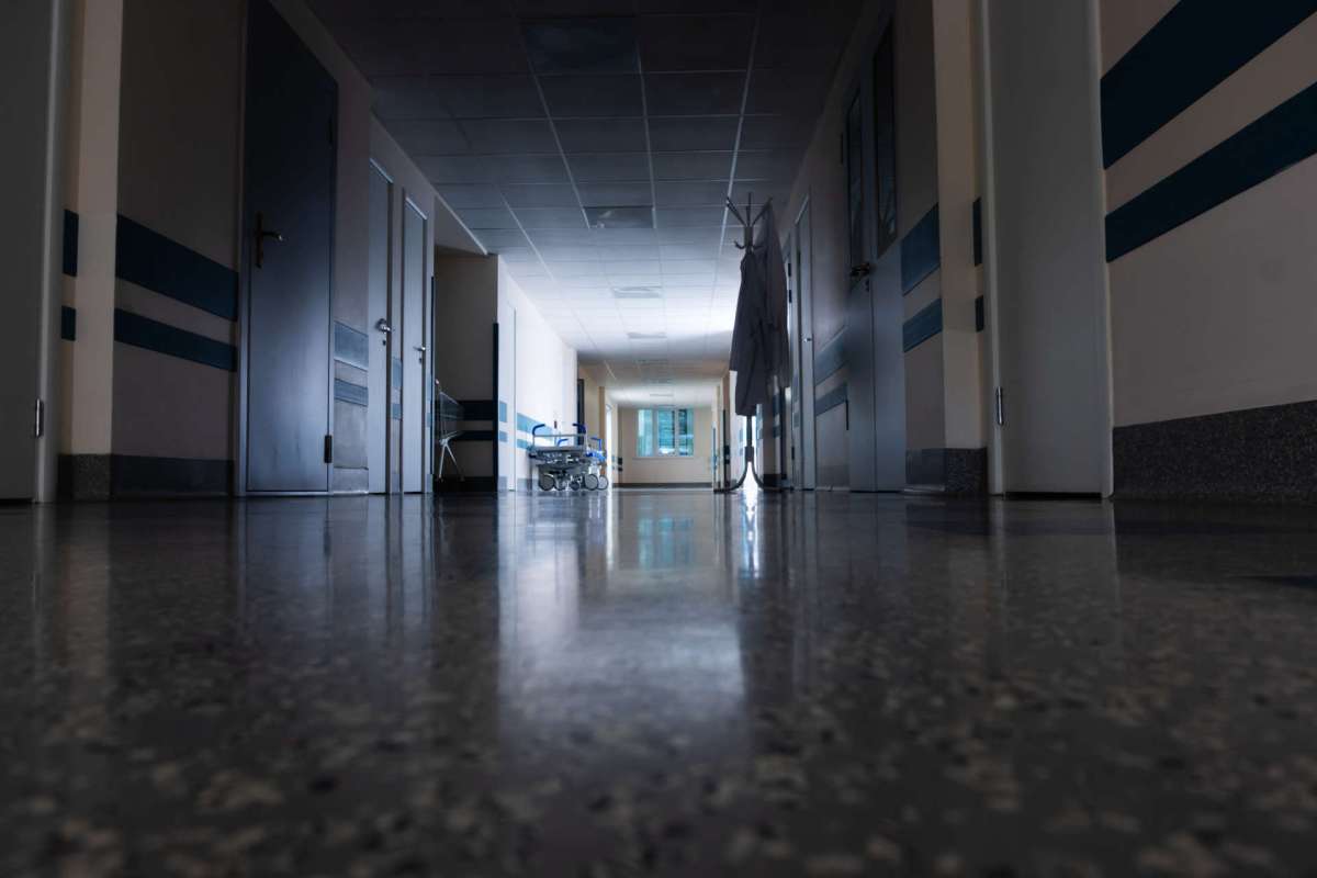 A corridor in a hospital with doors, a coat rack, and a bed visible in the background.