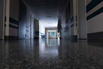 A corridor in a hospital with doors, a coat rack, and a bed visible in the background.