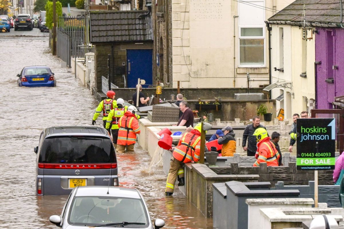 Emergency responders assist in a flooded street, with submerged vehicles and people trying to manage water on properties.