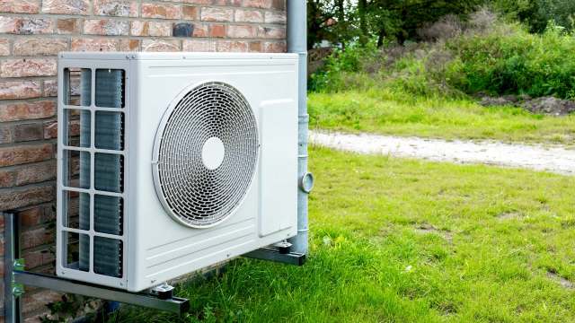 An air conditioning unit mounted outside a brick wall, next to a grassy area.