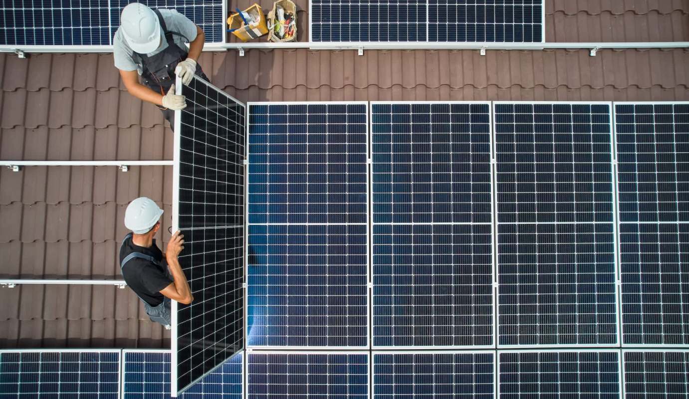 Two workers install solar panels on a roof with a box of supplies nearby.