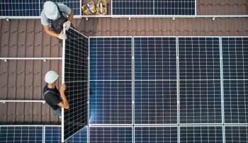 Two workers install solar panels on a roof with a box of supplies nearby.