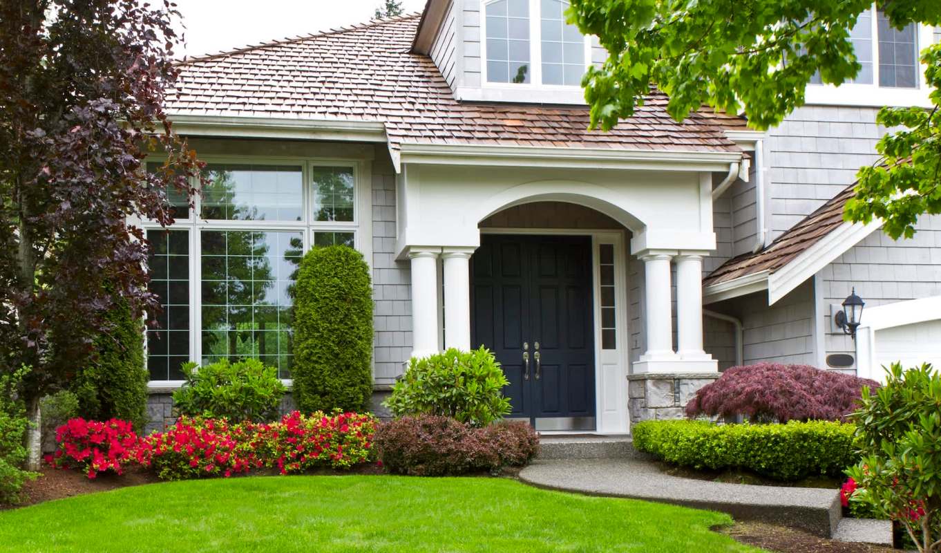 A house entrance featuring a porch, dark front door, and landscaped garden with colorful flowers and shrubs.