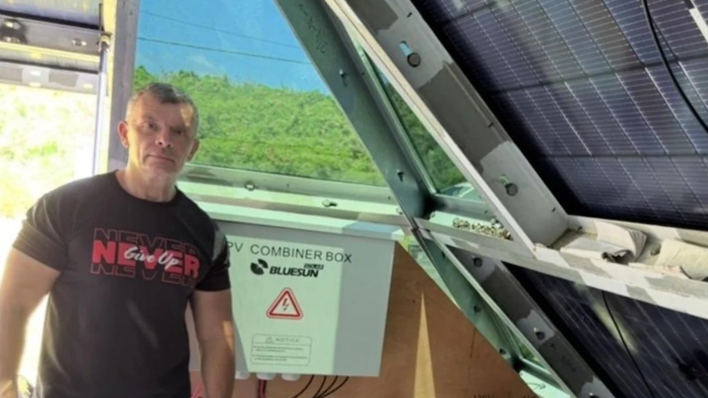 A person stands near a PV combiner box and solar panels, with greenery visible through an open structure.