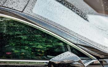 A Tesla car window covered in raindrops with greenery blurred in the background.