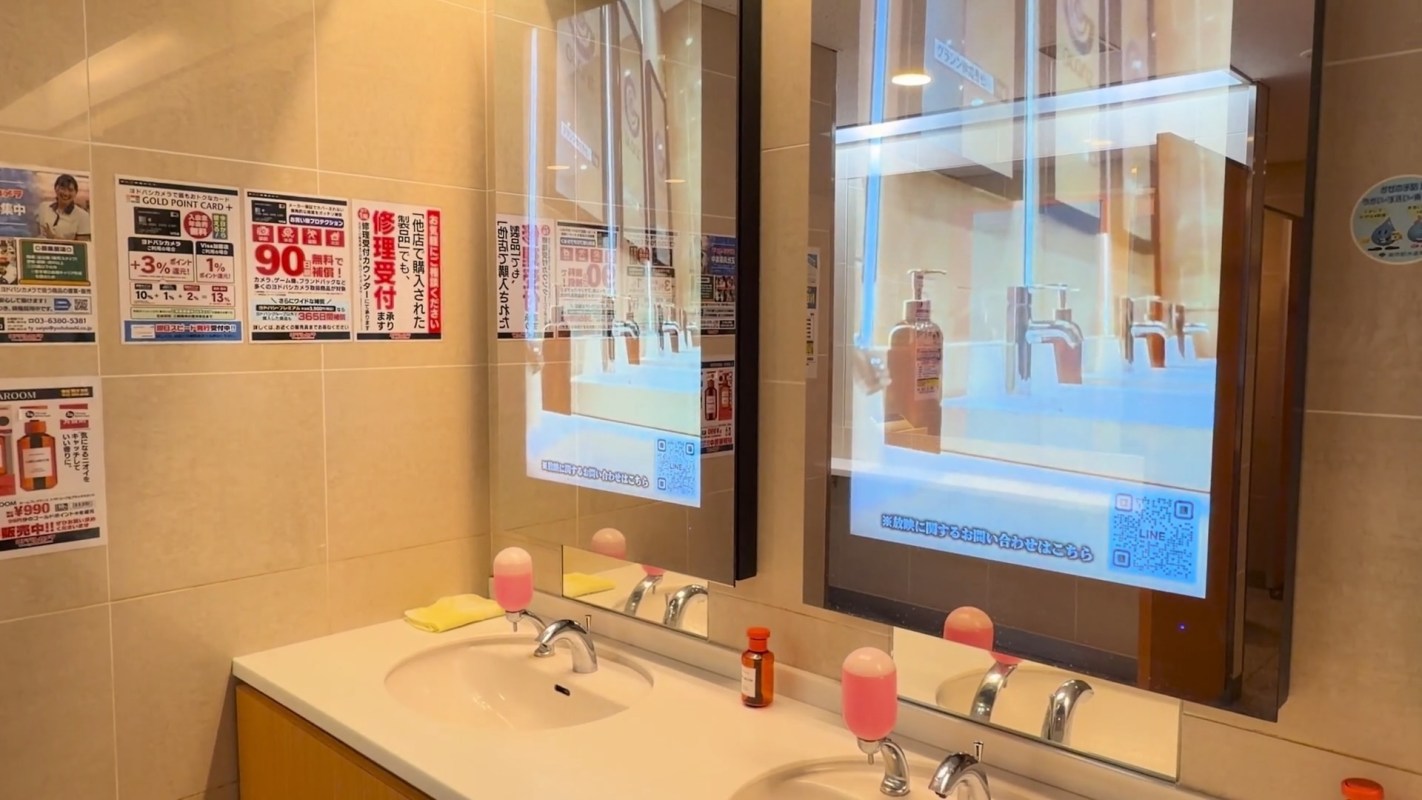 A bathroom with sinks, mirrors displaying advertisements, and pink liquid soap dispensers.
