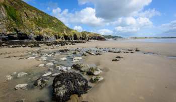 A sandy beach with rocks and seaweed, bordered by cliffs and a distant shoreline under a cloudy blue sky.